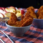 Two bowls filled with crispy breaded chicken tenders sit on a plaid cloth, with a plate of sliced apples and a fork and knife nearby. The background is softly lit.