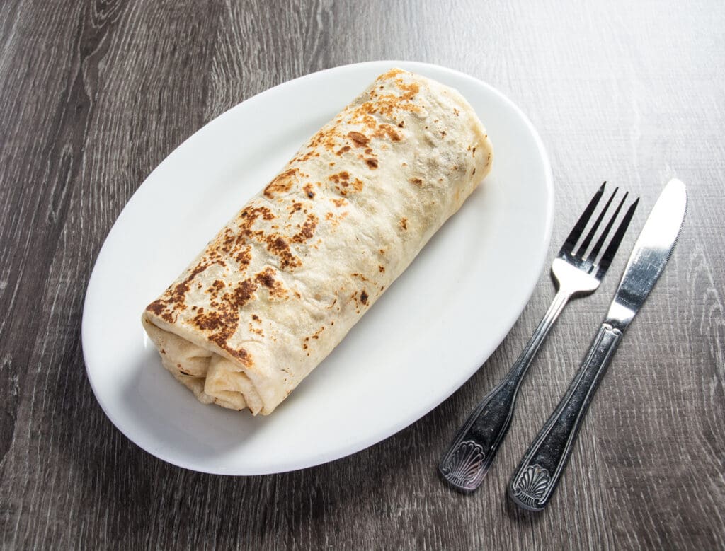 A large, grilled burrito wrapped in a tortilla sits on a white oval plate next to a fork and knife on a dark wooden table.