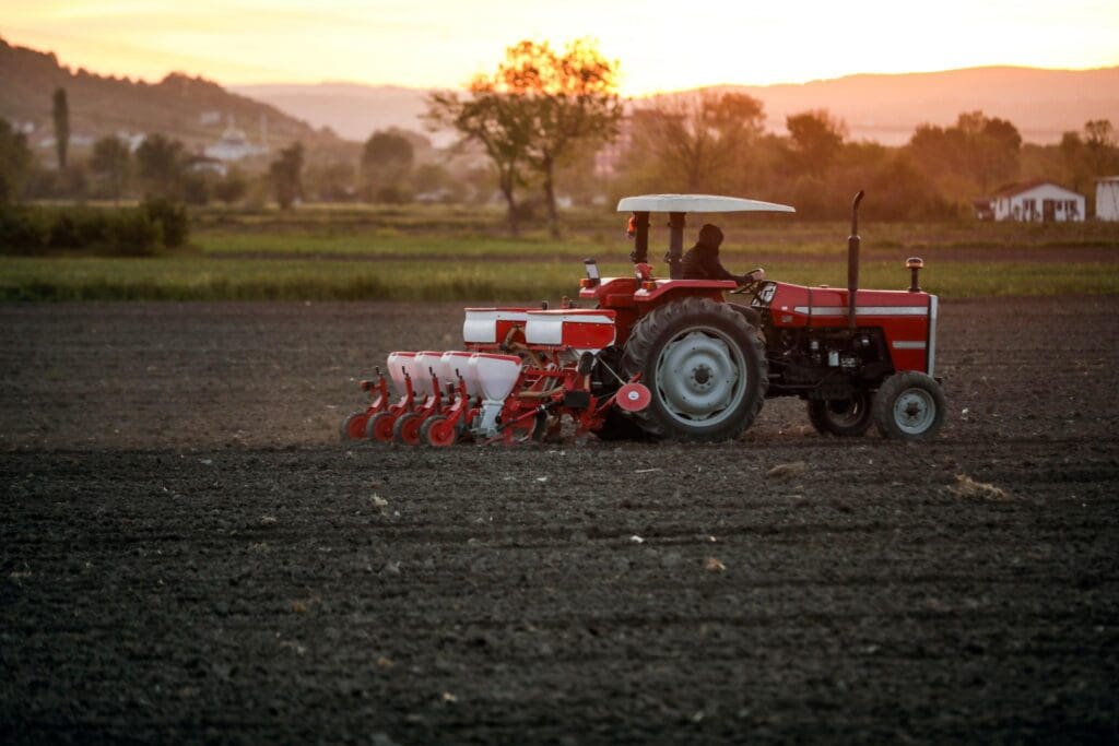 A farmer drives a red tractor with a seed planter attachment across a large, freshly plowed field at sunset, practicing sustainable farming with trees and distant buildings in the background.