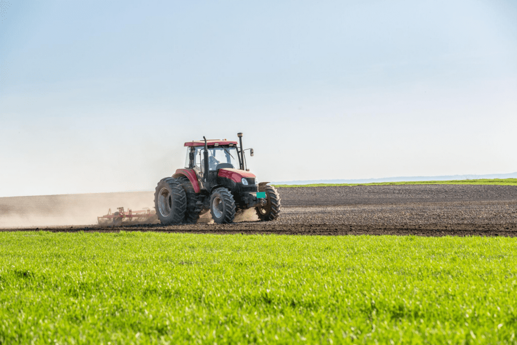 A red tractor plows a large, open field under a clear sky, illustrating the hard work and organic farming challenges faced in organic agriculture. Green grass lines the foreground while tilled soil stretches behind.