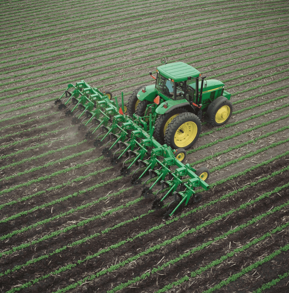 A green tractor with large yellow wheels pulls a multi-row cultivator through a field, tilling soil between rows of crops—a common scene in organic farming work. The striped pattern highlights the challenges and dedication of organic farming.