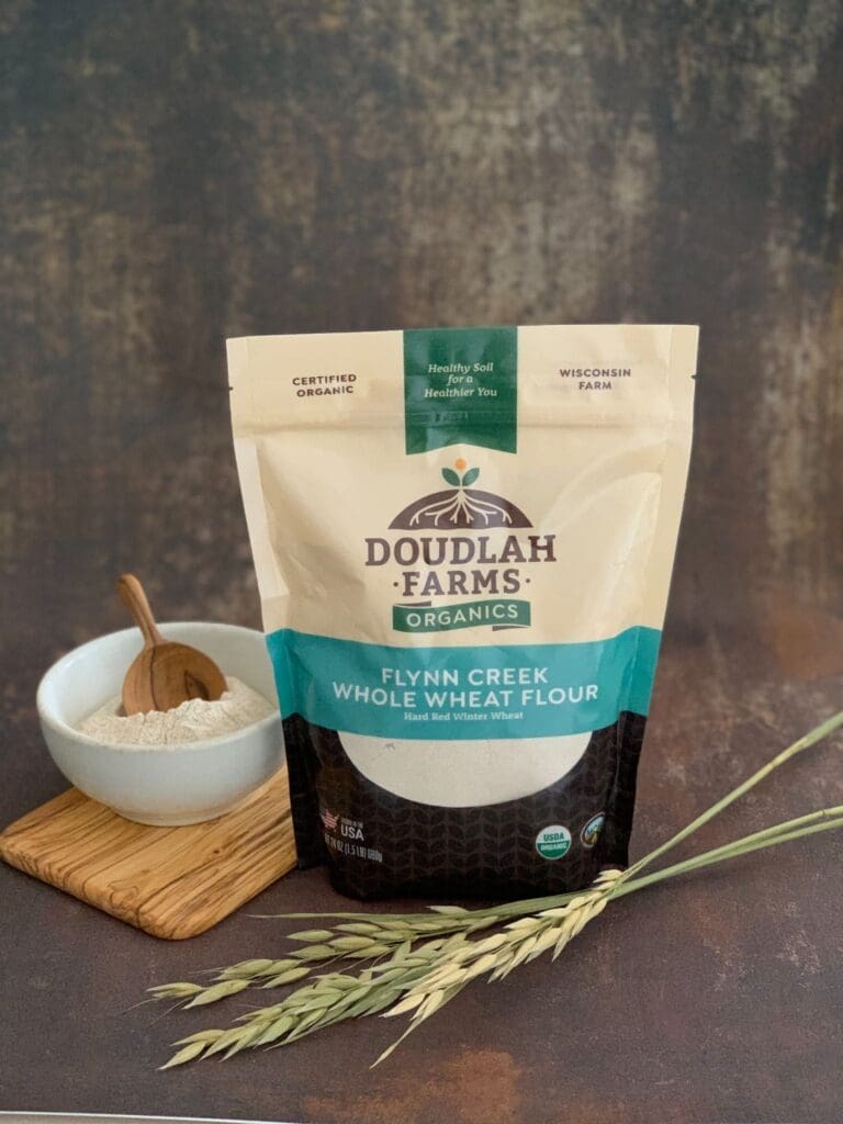 A bag of Doudlah Farms Organics Flynn Creek Whole Wheat Flour sits next to a bowl of flour with a wooden spoon, all on a wooden board with wheat stalks in front.