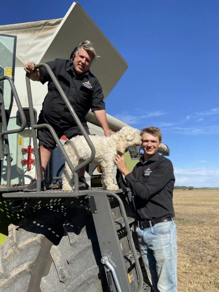 Two men in black shirts stand on the steps of farm equipment with a curly-haired white dog, smiling outdoors on a sunny day in a field with clear blue sky.